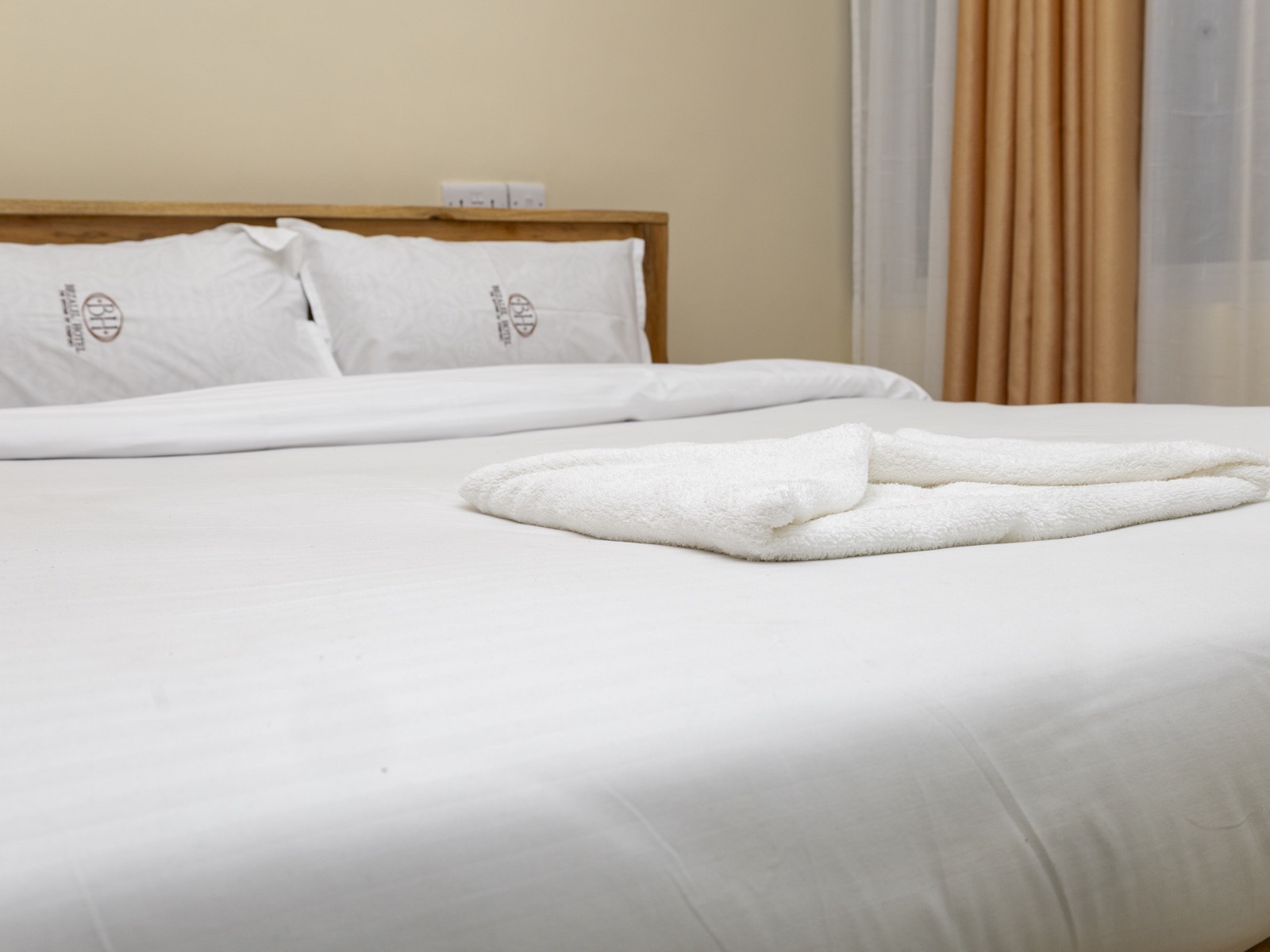 A made-up 5 by 6 bed with a folded towel, curtains drawn back